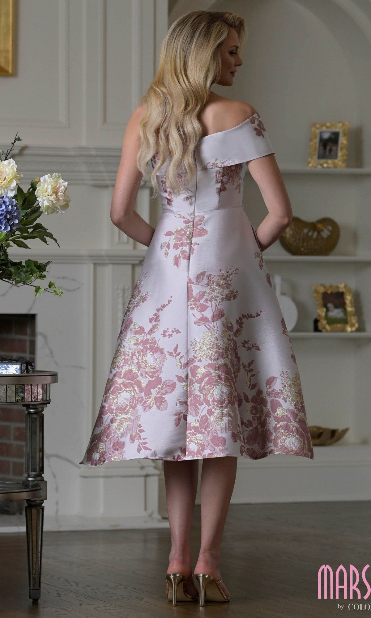 A woman stands indoors with her back to the camera, wearing the Marsoni MV1415 knee-length semi-formal dress, a floral off-the-shoulder fit-and-flare style paired with heels.
